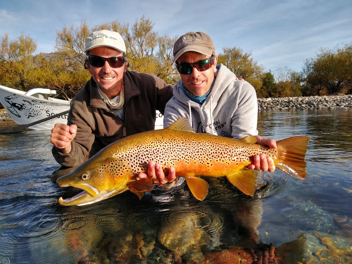 Dos pescadores sostienen una gran trucha marrón en el río Limay, con un bote y sauces de otoño detrás