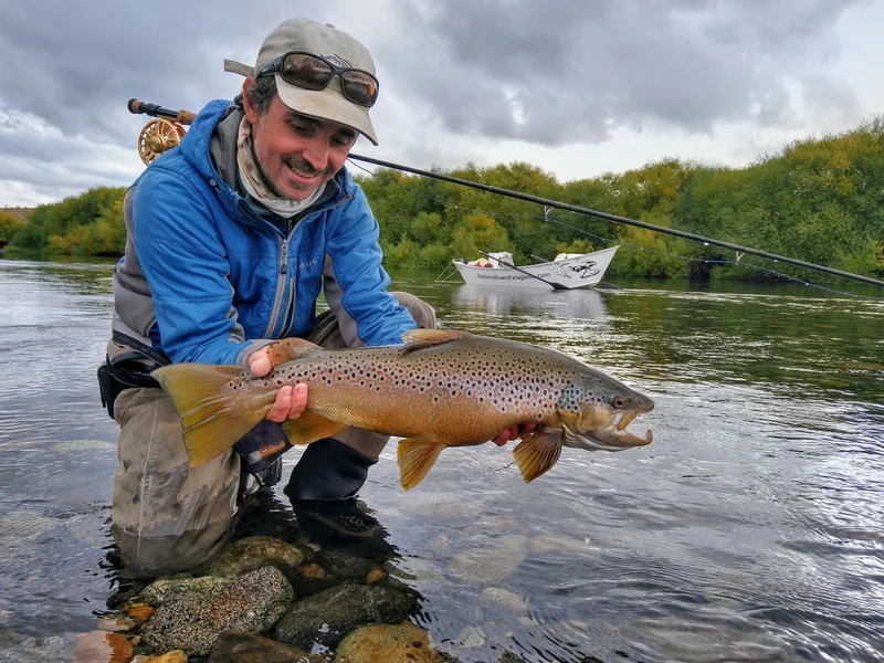 Pescador sosteniendo una trucha marrón junto a un bote durante una salida otoñal al Limay