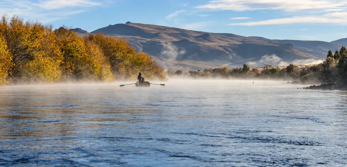Bruma de la mañana sobre el río Limay, con árboles y cerros reflejados en el agua