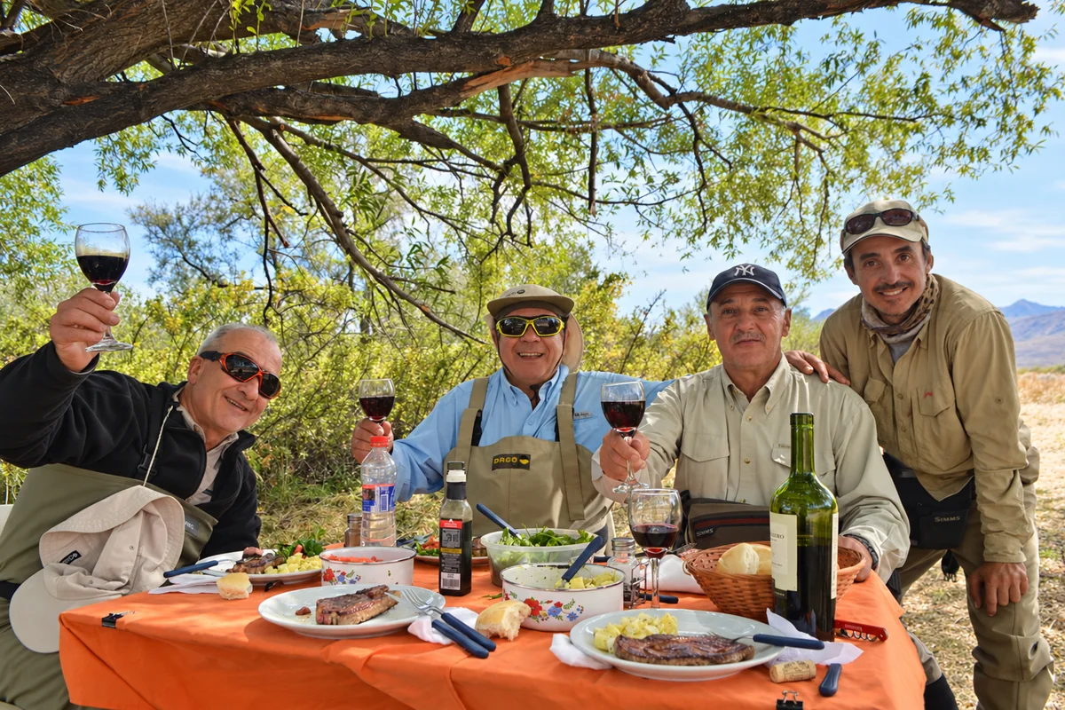 Un grupo de pescadores comparte el almuerzo y brinda con copas de vino en una mesa junto al río Manso