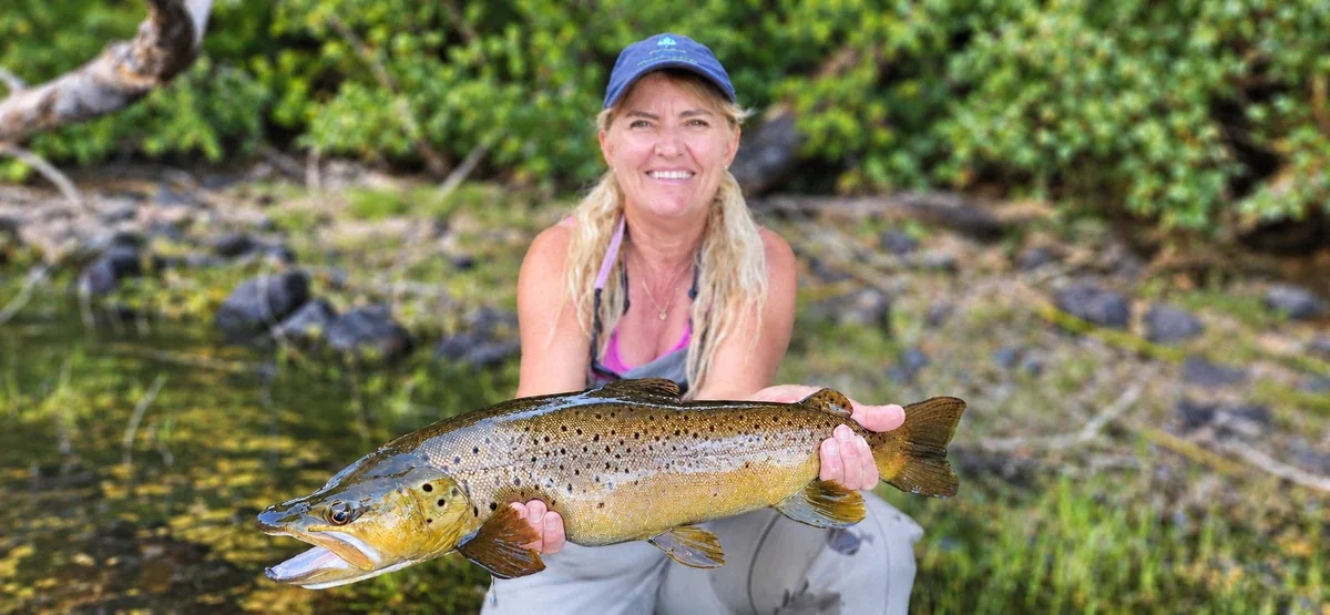 Pescadora sosteniendo una trucha marrón junto a un lago boscoso de Patagonia
