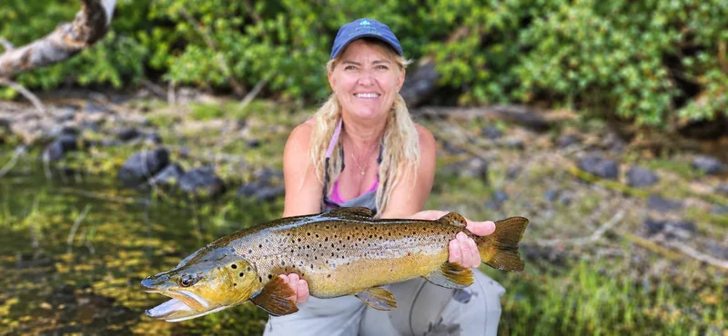 Pescadora sosteniendo una trucha marrón junto a un lago boscoso de Patagonia