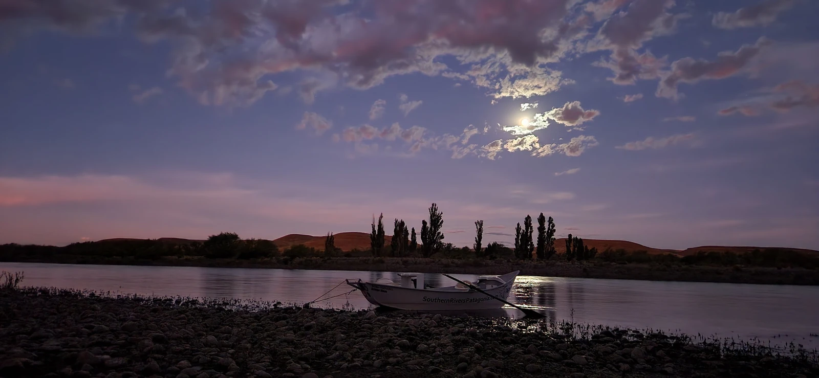 Drift boat moored on the Collón Cura River under the moonlight during an overnight fly fishing expedition in Patagonia.
