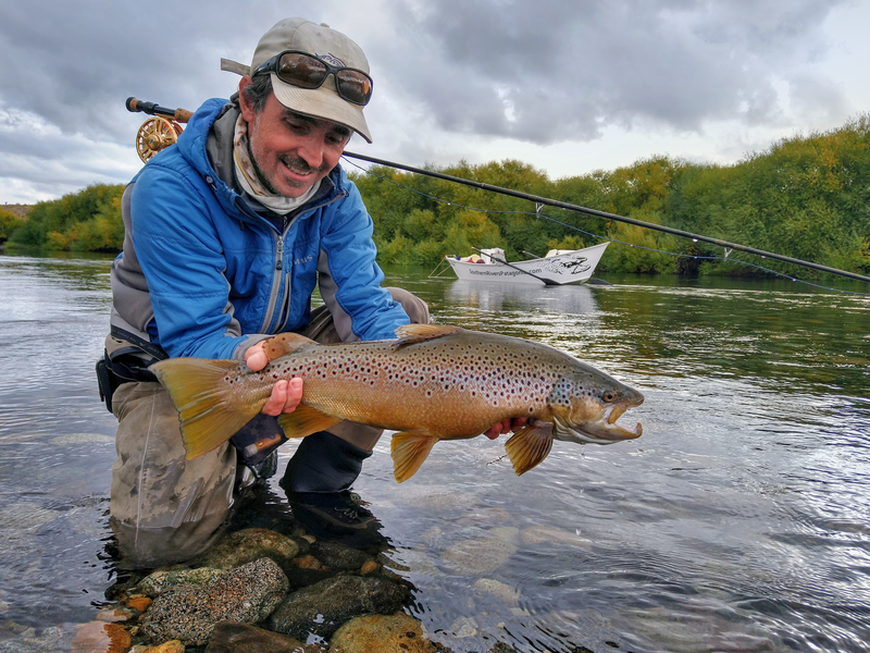 Pescador con una trucha marrón en el río Limay, con una embarcación de Southern Rivers Patagonia al fondo