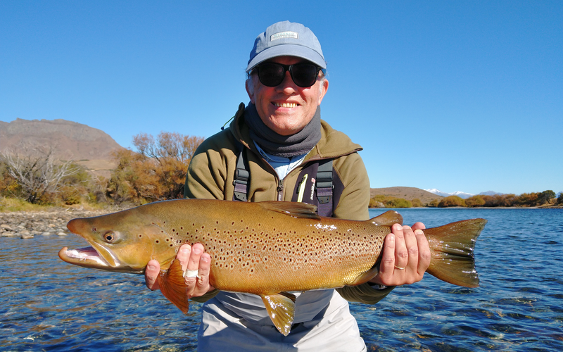 Pescador con una trucha marrón en el río Limay, cerca de Bariloche, con árboles de otoño en las orillas