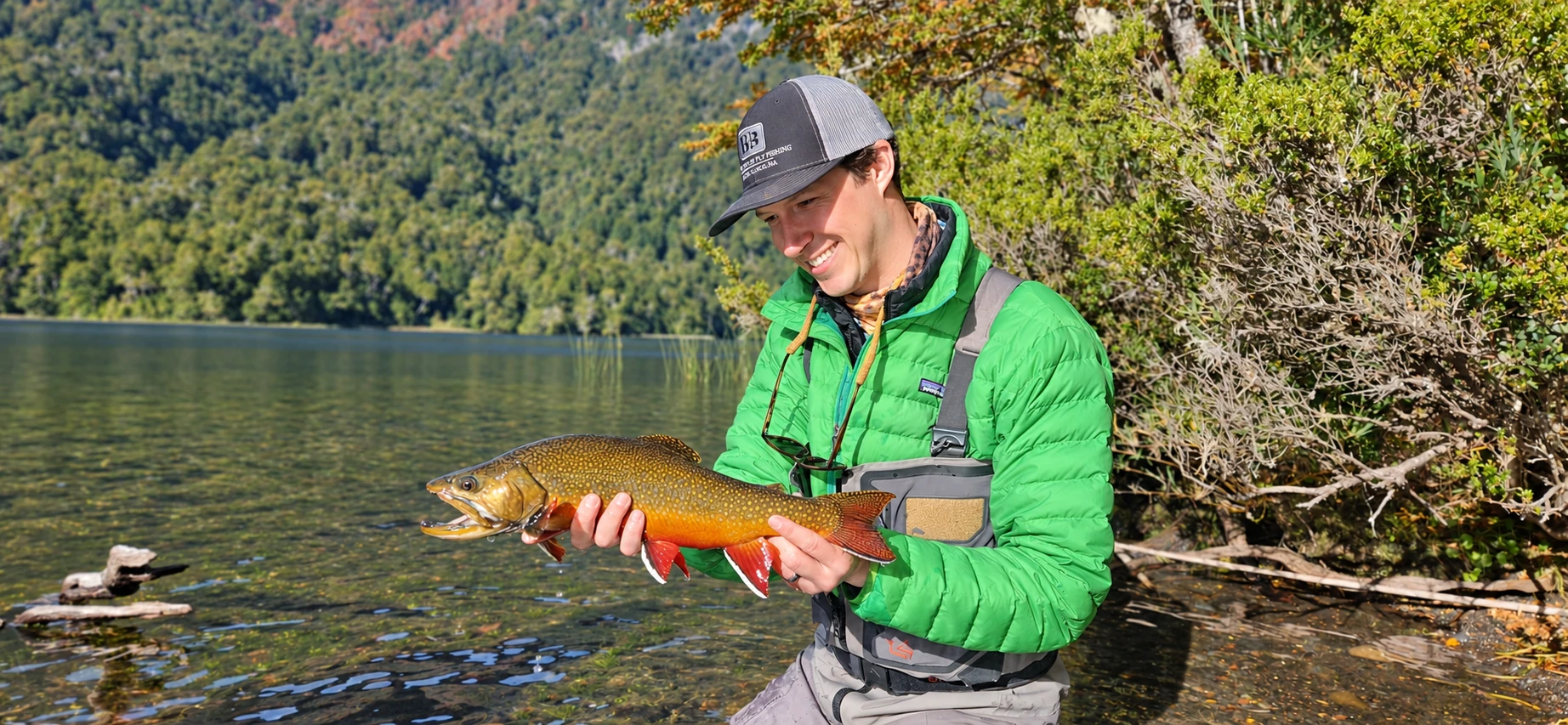 Pescador sosteniendo una trucha fontinalis en la orilla del lago Fonck, rodeado de bosque nativo, cerca de Bariloche, Patagonia Argentina.